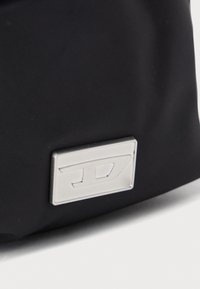 Svart nylonväska med en silverfärgad rektangulär logoplåt med präglad text. Slät textur, minimalistisk design, lämplig för olika användningsområden.