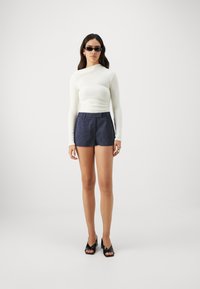 Top de manga larga crema, ajustado en la cintura; pantalones cortos azul oscuro a rayas; sandalias de tacón negras; gafas de sol rectangulares negras.