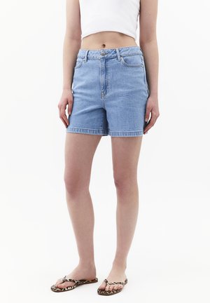 Person trägt hellblaue Jeansshorts, weißes kurzes Top und Leopardenmuster-Flip-Flops, steht mit entspannten Händen an den Seiten.