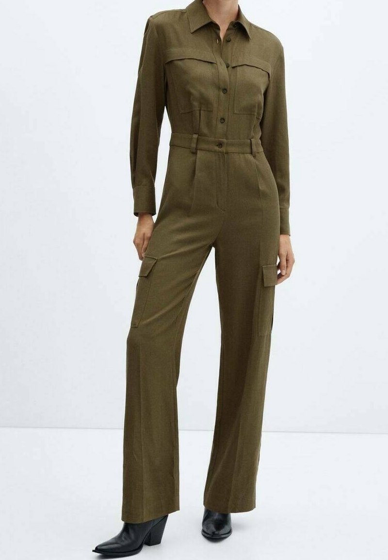 Femme portant une combinaison vert olive boutonnée avec poches à l'avant et pantalon à jambes larges, associée à des bottines noires, debout sur fond blanc.