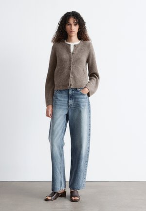 Ženska z kodrastimi lasmi, oblečena v rjavo gumbasto jopico, belo srajco, ohlapne modre kavbojke in rjave sandale z odprtimi prsti, stoji na sivi tleh.