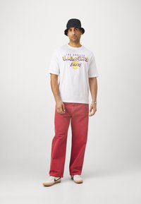 Bijela majica s ljubičastim i žutim logom "Los Angeles Lakers", u kombinaciji s crvenim hlačama i crnom kbelj šapkom. Casual obuća s bijelim i crnim detaljima.