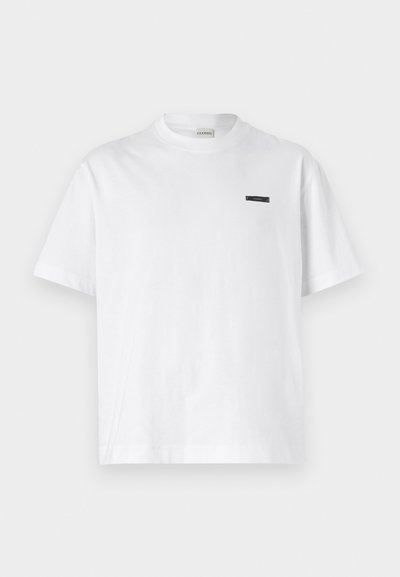 Balta kokvilnas T-kreklā ar apaļu kakla izgriezumu. Tam ir neliela melna birka uz krūtīm. Īsas piedurknes un brīva piegriezuma dizains.