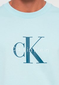 Krupan kadar svijetloplave košulje s velikim tamnoplavim slovima "CK" i tekstom "Calvin Klein" izvezenim na prsima.