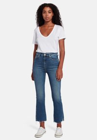 T-shirt bianco con scollatura a V, jeans blu a vita alta con orlo sfilacciato, modello che indossa sneakers bianche, tessuto morbido, vestibilità casual.