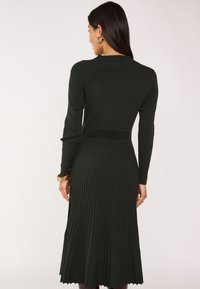 Abito a maglia rib knit verde scuro con maniche lunghe, corpino aderente e gonna svasata; presenta un orlo scallop e linee verticali testurizzate.