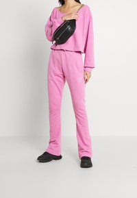 Roze sweatshirt en bijpassende joggingbroek van zacht materiaal, met een losse pasvorm en logo-accenten. Draagt met een zwarte schoudertas en zwarte schoenen.