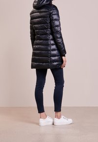 Abrigo acolchado negro con capucha, con acolchado horizontal, corte de longitud media, combinado con vaqueros oscuros y zapatillas blancas.