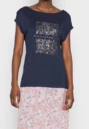 Personne portant un t-shirt bleu marine avec des carrés floraux argentés métalliques et un texte, associé à une jupe blanche ornée de points roses, rouges et noirs.