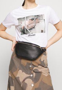 Hvid t-shirt med et trykt billede og teksten "Pivot!" parret med en sort læder bæltetaske med guld lynlås og buet form.