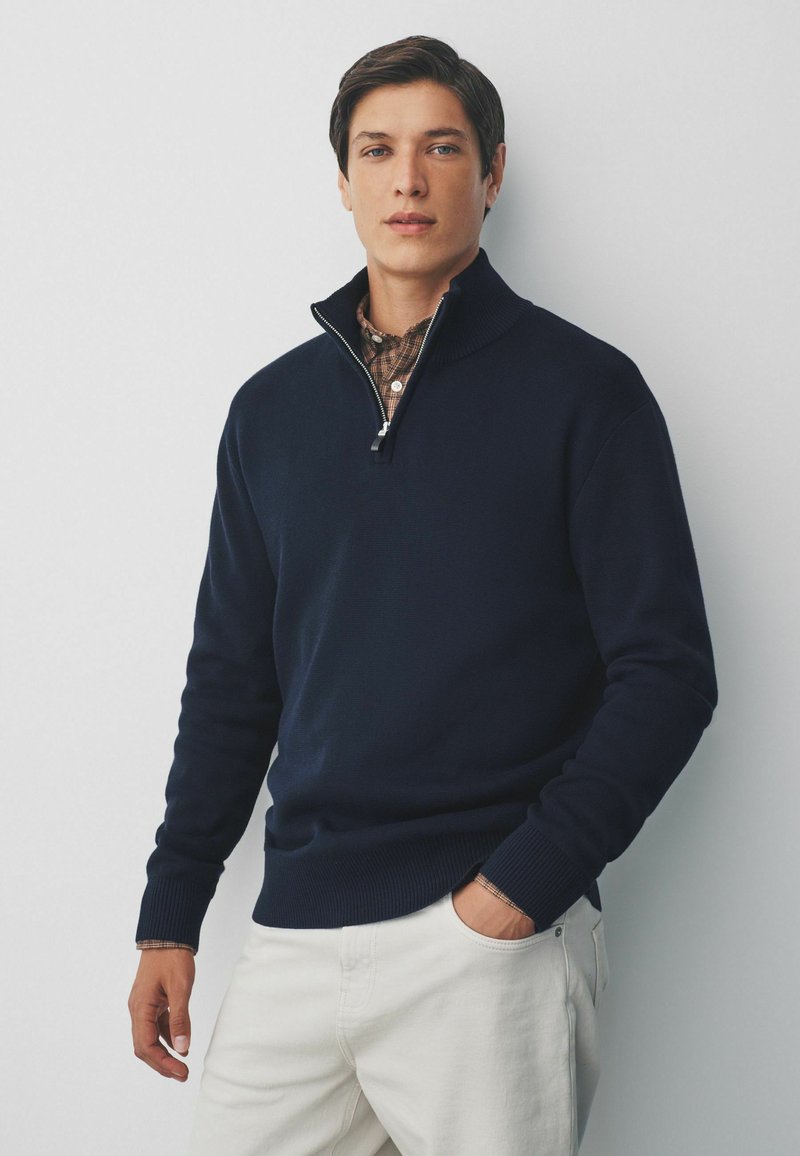 Marineblauer Pullover mit Reißverschluss aus Strickstoff, mit geripptem Saum und Bündchen, kombiniert mit einem karierten Hemd darunter.
