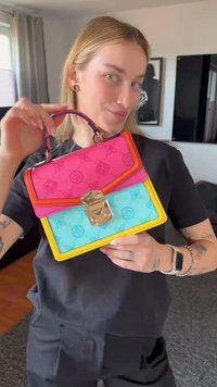 Femme tenant un petit sac à main avec des panneaux roses, bleus et jaunes et une fermeture dorée, debout dans une pièce avec une télévision et des œuvres encadrées.