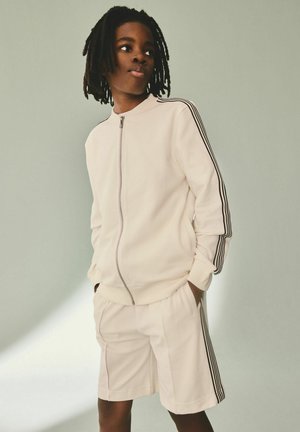 Giovane con dreadlocks che indossa una giacca bianca con cerniera e pantaloncini abbinati con strisce nere sulle maniche e sui lati, che guarda di lato.