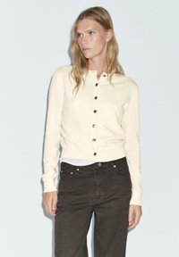 Cardigan en laine crème avec un col rond, des boutons décoratifs sur le devant et des manches longues, associé à un jean marron foncé.