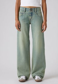 Άτομο που φοράει ανοιχτό μπλε τζιν παντελόνι με φαρδιά μπατζάκια και διπλό κούμπωμα με κουμπιά, λευκό κοντό τοπ και λευκά αθλητικά παπούτσια με καφέ σόλες.