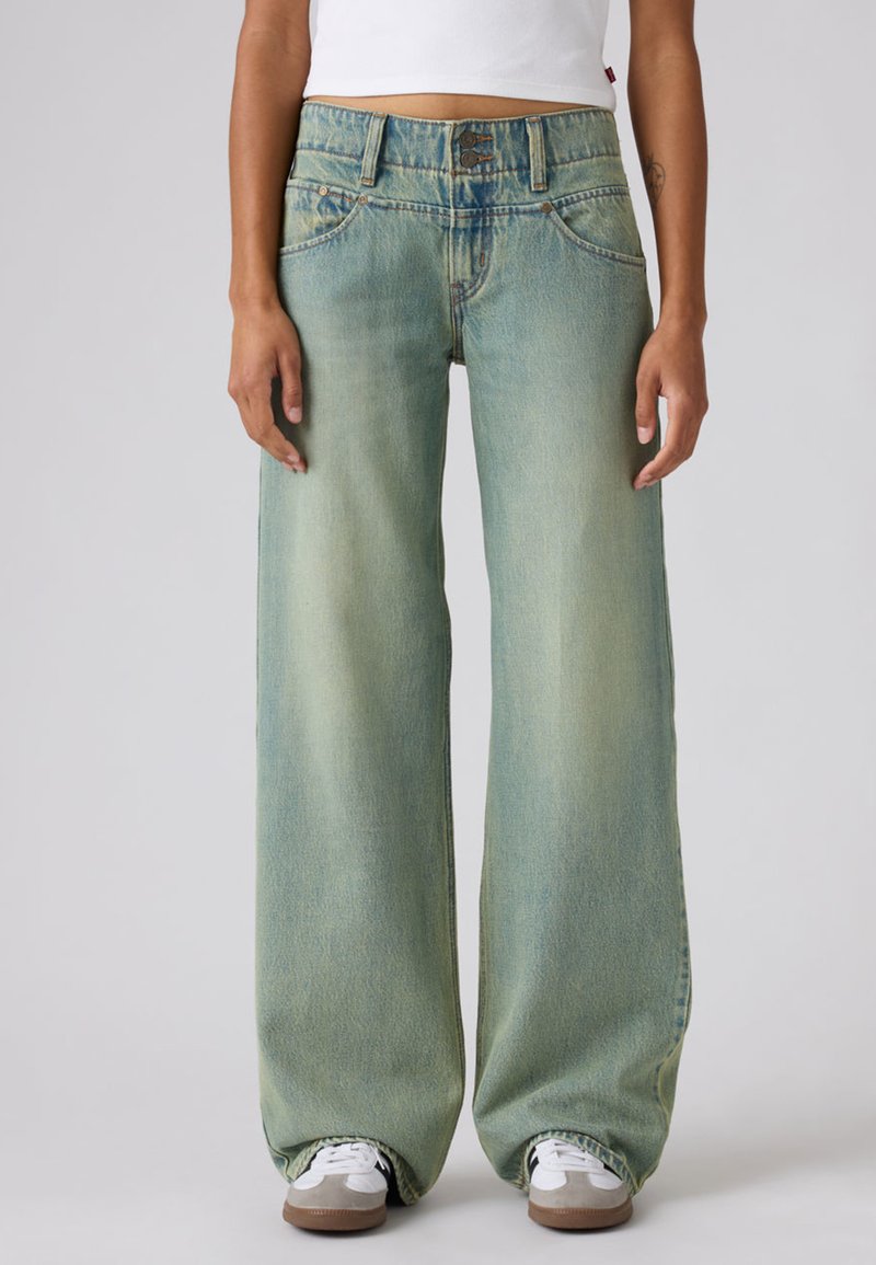 Άτομο που φοράει ανοιχτό μπλε τζιν παντελόνι με φαρδιά μπατζάκια και διπλό κούμπωμα με κουμπιά, λευκό κοντό τοπ και λευκά αθλητικά παπούτσια με καφέ σόλες.