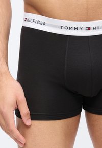 Άνδρας που φοράει μαύρα εσώρουχα Tommy Hilfiger με λευκή μέση που φαίνεται το όνομα και το λογότυπο της μάρκας, το χέρι ακουμπάει στο μηρό.