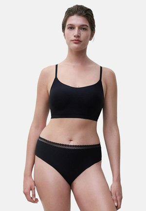 Woman wearing a black seamless bralette and black high-cut underwear with lace trim, standing against a plain light gray background.