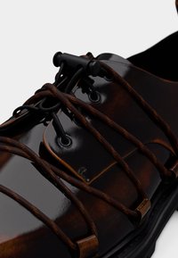 Γυαλιστερό, σκοτεινό καφέ παπούτσι με μαύρα και καφέ ελαστικά κορδόνια χωρίς δέσιμο, ασφαλισμένα με μαύρο διακόπτη κοντά στη γλώσσα.