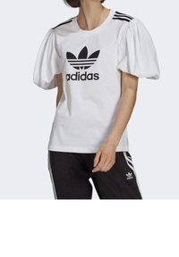 Camiseta blanca con mangas abullonadas, que presenta un logo de Adidas en negro y tres rayas negras en los hombros. Combinada con pantalones negros.