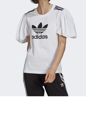 Camiseta blanca con mangas abullonadas, que presenta un logo de Adidas en negro y tres rayas negras en los hombros. Combinada con pantalones negros.
