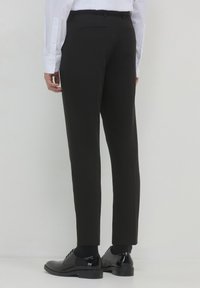 Persona con pantaloni neri su misura, scarpe eleganti nere e una camicia bianca con bottoni, in piedi contro uno sfondo chiaro.