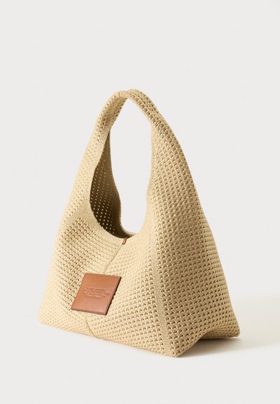 Sac cabas tissé beige avec des anses larges et une petite pièce rectangulaire en cuir marron sur le devant.