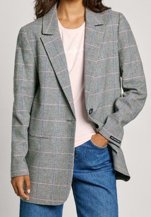 Kobieta w szarej kratkowanej marynarce na jasnoróżowej koszulce i niebieskich dżinsach, z jedną ręką w kieszeni, stojąca na jednolitym tle.