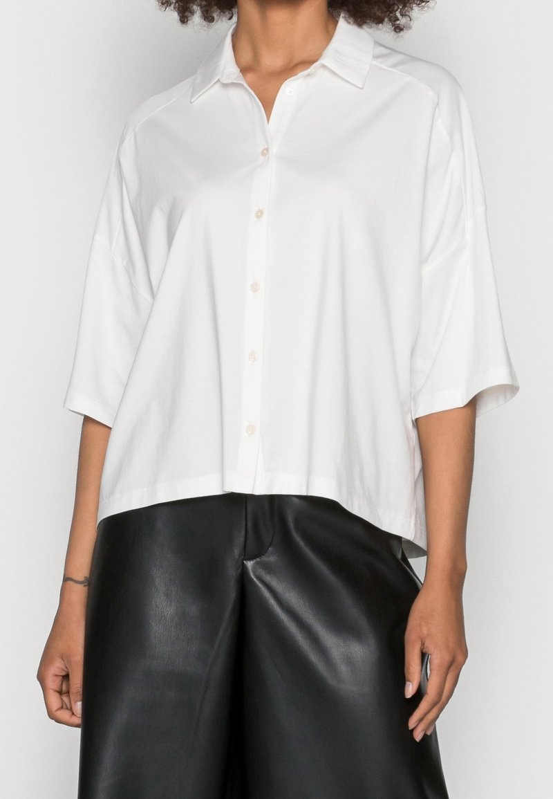 Femme aux cheveux bouclés portant une chemise blanche ample à boutons avec des manches trois-quarts et un pantalon large en cuir noir.