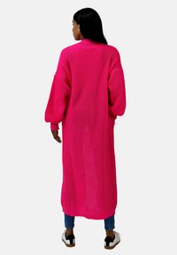 Cardigan tricoté fuchsia, long et surdimensionné, avec un motif texturé et de larges manches, porté avec un jean bleu et des baskets noires et blanches.