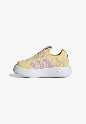 Zapatilla deportiva de punto amarilla tipo slip-on con rayas lila claro, suela blanca gruesa y pestañas en el talón y la lengüeta sobre un fondo blanco.