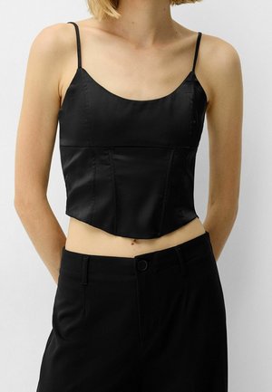 Kvinde iført sort satin cropped camisole med tynde stropper og sort højtaljede bukser mod en ensfarvet baggrund.