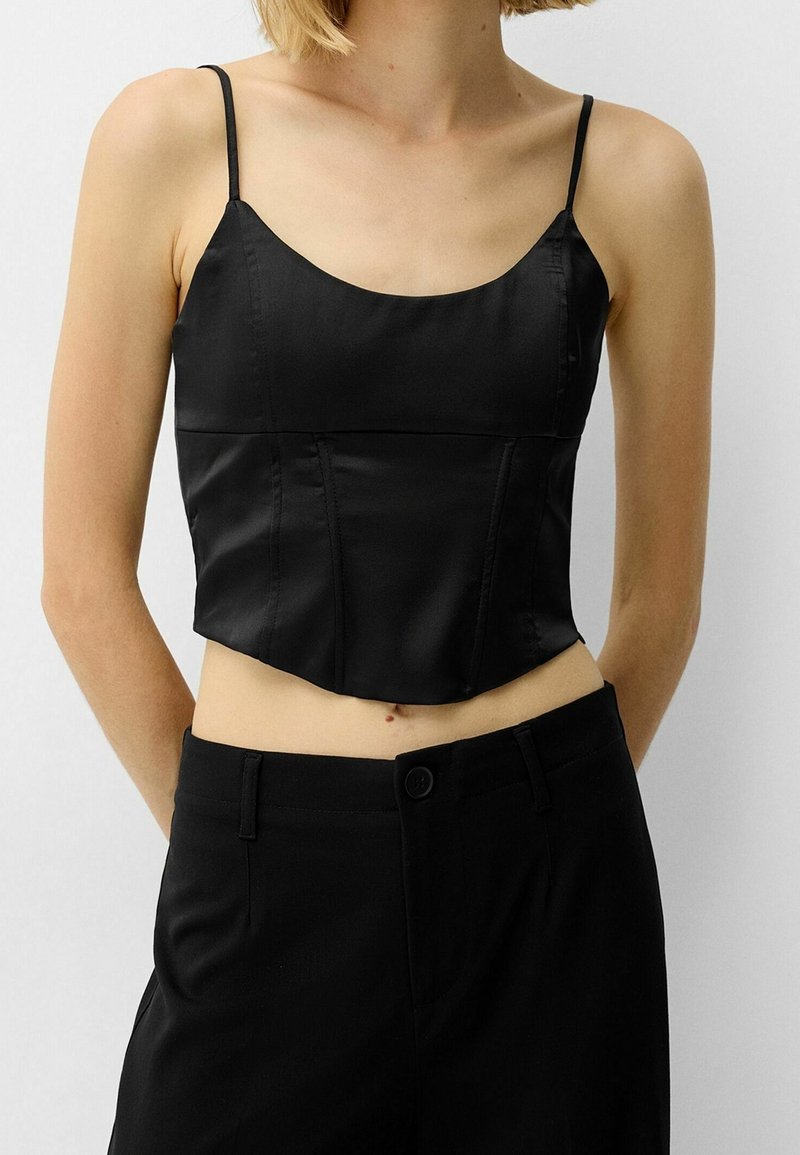 Femme portant un caraco court en satin noir à fines bretelles et un pantalon noir taille haute sur un fond uni.