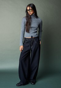 Sivý rebrovaný rolák v kombinácii so širokými tmavými džínsami. Model nosí priehľadné slnečné okuliare; pozadie je jemne zelené.