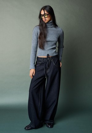 Sivý rebrovaný rolák v kombinácii so širokými tmavými džínsami. Model nosí priehľadné slnečné okuliare; pozadie je jemne zelené.