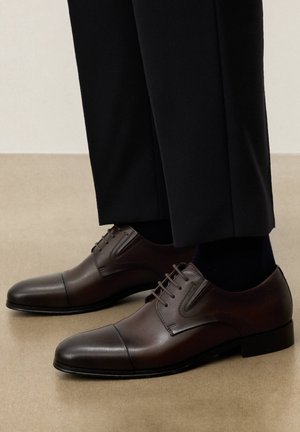 Rjave usnjene klasične čevlje s kapico, nošene z črnimi hlačami in temnimi nogavicami na bež površini tal.