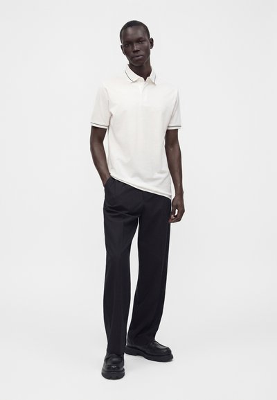 Homme portant un polo blanc à manches courtes, un pantalon noir et des chaussures noires, debout devant un fond blanc uni.
