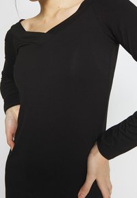 Svart långärmad topp med en unik V-formad halsringning och figurnära design; tillverkad av ett mjukt tyg med en lätt glans och stretch.