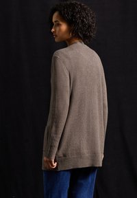 Brązowy kardigan o teksturze z prążkowanymi mankietami i dołem, z bocznymi kieszeniami i luźnym krojem, w połączeniu z niebieskimi spodniami dżinsowymi.