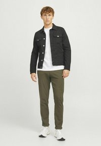 Jachetă din denim neagră cu nasturi argintii, tricou alb, pantaloni verzi măslinii și adidași albi. Modelul stă în picioare pe un fundal simplu.