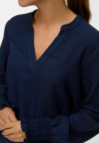 Donna che indossa una blusa lunga a maniche lunghe di un blu navy con scollo a V e polsini arricciati, mani delicate unite in vita.