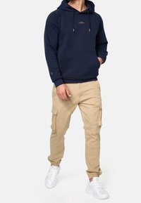 Marineblå hoodie med kengurulomme og snore, parret med beige cargo bukser med sidelommer, afsluttet med hvide sneakers.