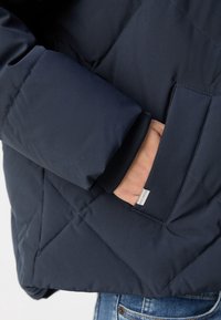 Veste matelassée bleu marine avec une poche latérale, des poignets côtelés et une texture lisse. Étiquette accent visible près de la poche.