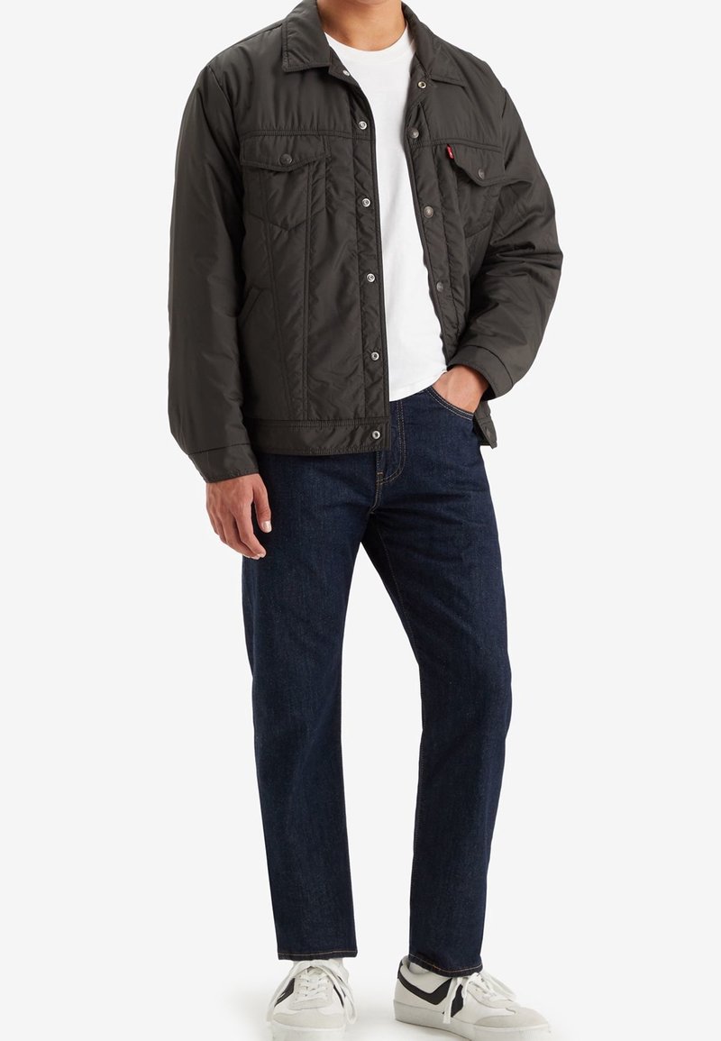 Homme portant une veste matelassée noire sur une chemise blanche, un jean bleu foncé et des baskets blanches, debout avec une main dans la poche.