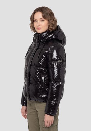 Une femme porte une doudoune noire brillante à capuche et un pantalon cargo olive sur un fond clair et uni.
