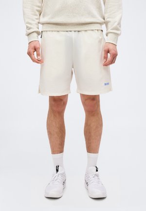 Mężczyzna ubrany w kremowe spodenki sportowe, białe skarpetki za kostkę oraz białe trampki, stojący na jednolitym jasnym tle.