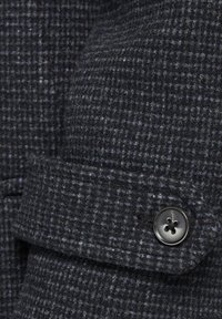 Mörk mönstrad textil med en subtil rutdesign. Har en svart knapp med fyra hål och en vikbar manschettdetalj. Mjuk textur.