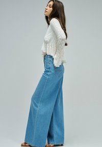 Mujer con cabello largo y castaño, vestida con una blusa blanca de encaje de manga larga y jeans azules de tiro alto y pierna ancha, de pie de perfil.