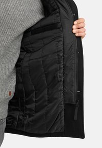 Schwarze gepolsterte Jacke mit einem glatten, abgesteppten Innenfutter, ausgestattet mit einem Reißverschluss und einer Innentasche. Das äußere Gewebe hat eine weiche Textur.