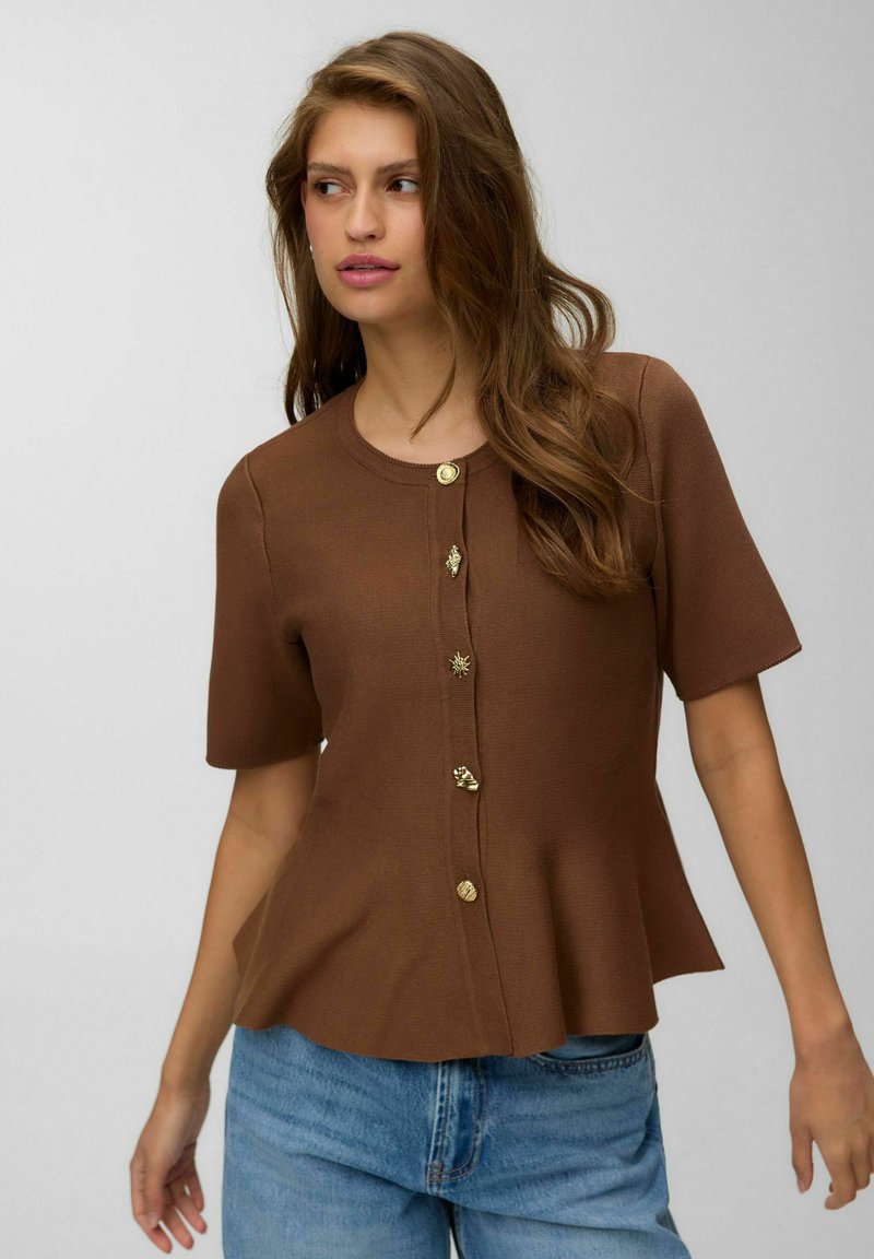 Femme portant un cardigan marron à manches courtes avec des boutons dorés uniques, associé à un jean bleu clair, regardant sur le côté.